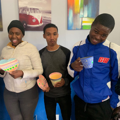 A group of participants from a local charity proudly holding their newly painted pottery pieces, created through Clay Café’s Paint and Donate initiative, supporting pottery painting for local charities.